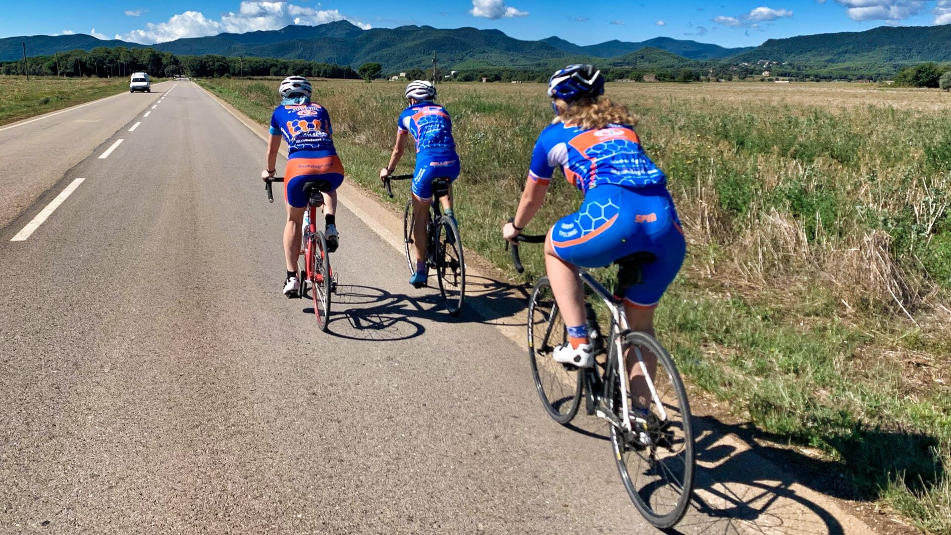 Three cyclists in matching blue kits ride on a sunny country road near Girona, Spain, with hills ahead