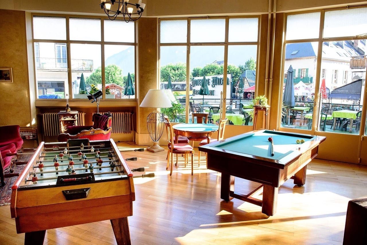Bright game room at Sport'Hotel-Aparthotel de Milan with foosball, pool table, wooden floor and large windows showing outdoor seating