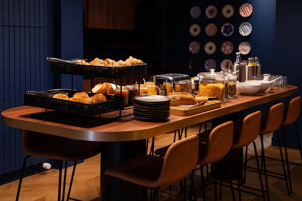 Breakfast buffet setup with croissants, bread, cereals, jams, and plates on a wooden table with brown chairs in a cozy dining area