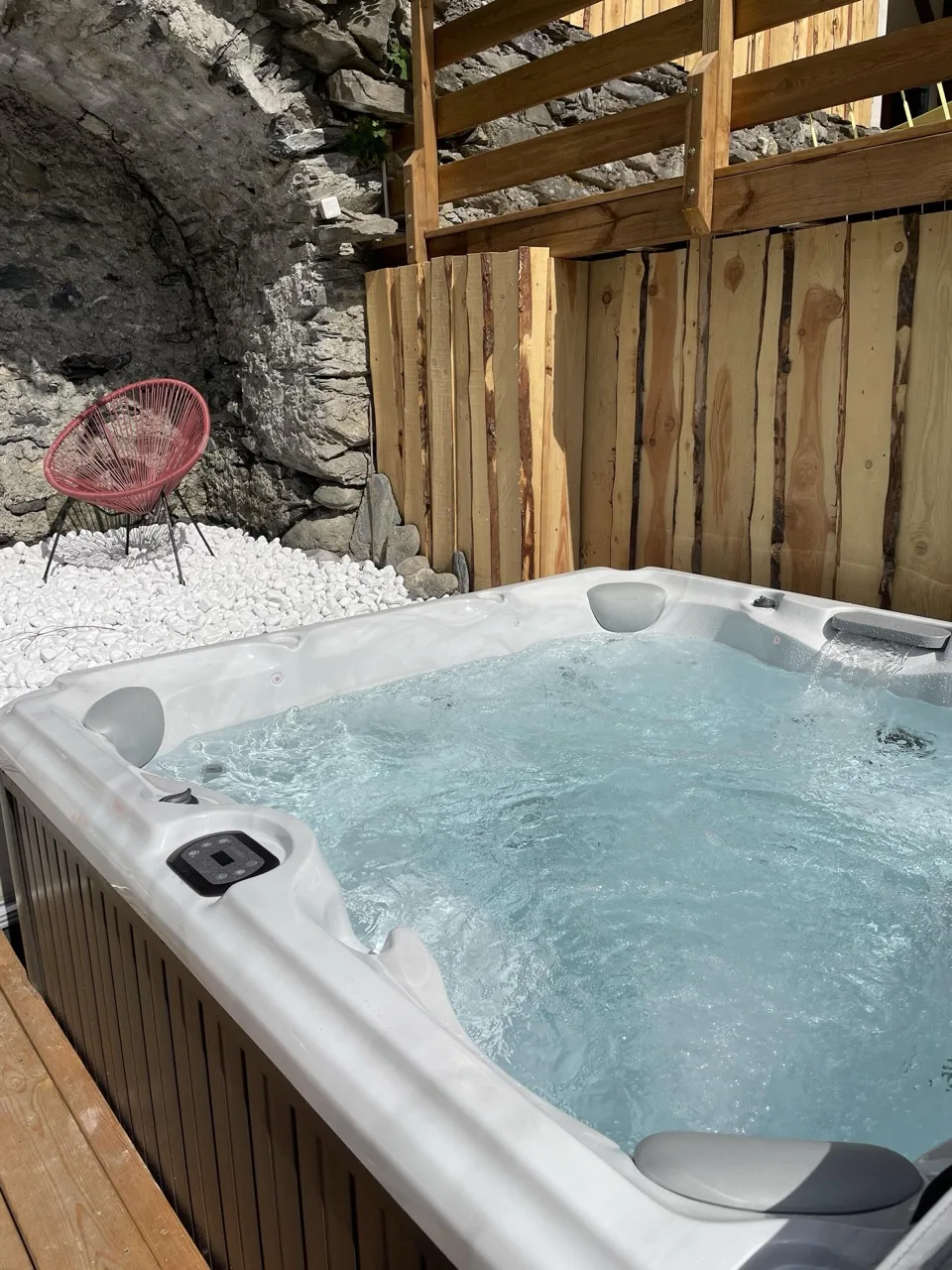 Large jacuzzi with bubbling water set next to a wooden fence and rustic stone wall, with white pebbles and a red chair nearby