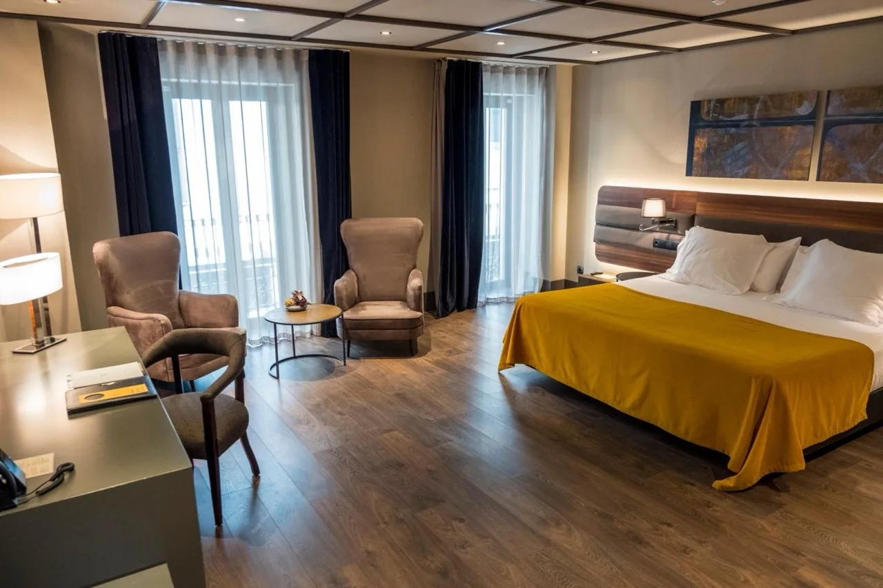 Spacious bedroom at Hotel Nord 1901 in Girona with light brown walls, brown furniture, and white bed with mustard throw