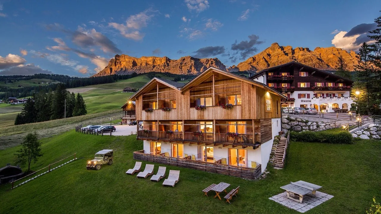 Distant view of Hotel Rezia in La Villa with mountain backdrop, greenery, and sun loungers in front
