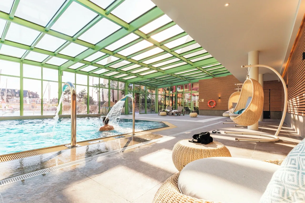 Indoor pool at Résidence Pierre & Vacances Premium Presqu'Ile de la Touques Deauville with glass ceiling, walls, and stylish chairs