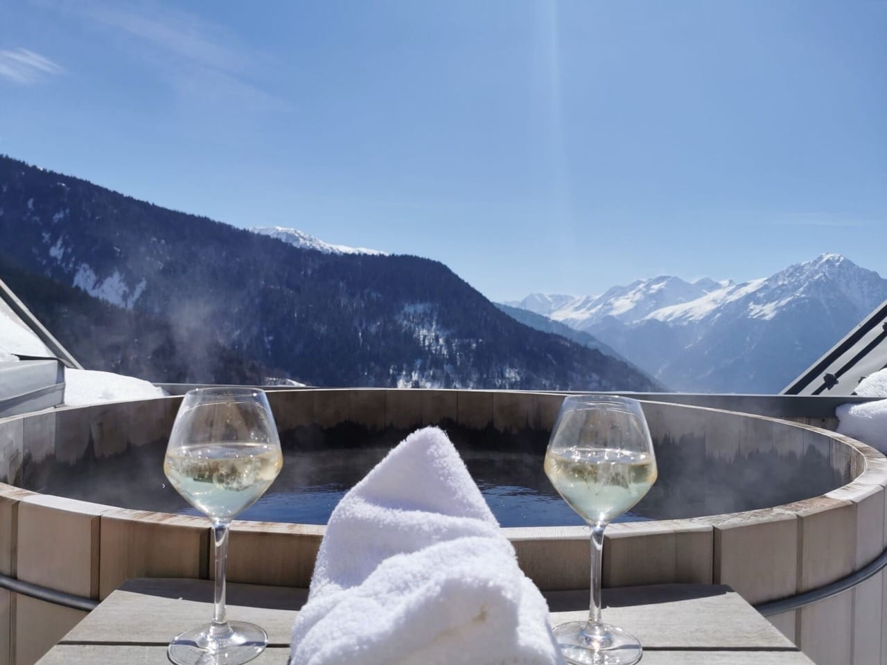 Rooftop jacuzzi with rustic brick design and two wine glasses overlooking mountains at Hotel Le V de Vaujany