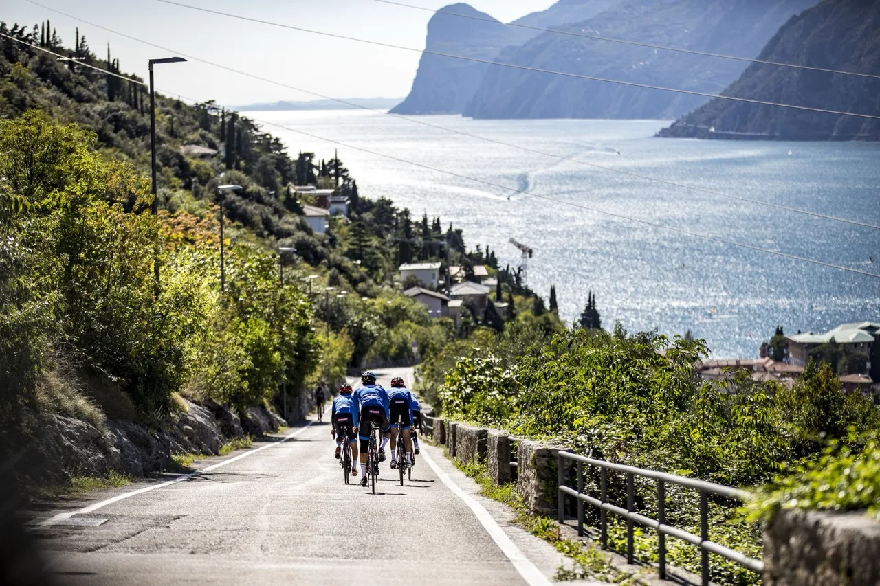 Three cyclists riding on a road with the sea and mountains in the background under a clear sky