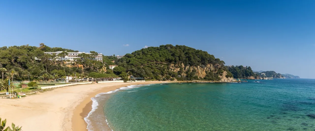 "Beach view at Hotel Santa Marta in Lloret de Mar, showing golden sand, calm sea, and lush greenery nearby