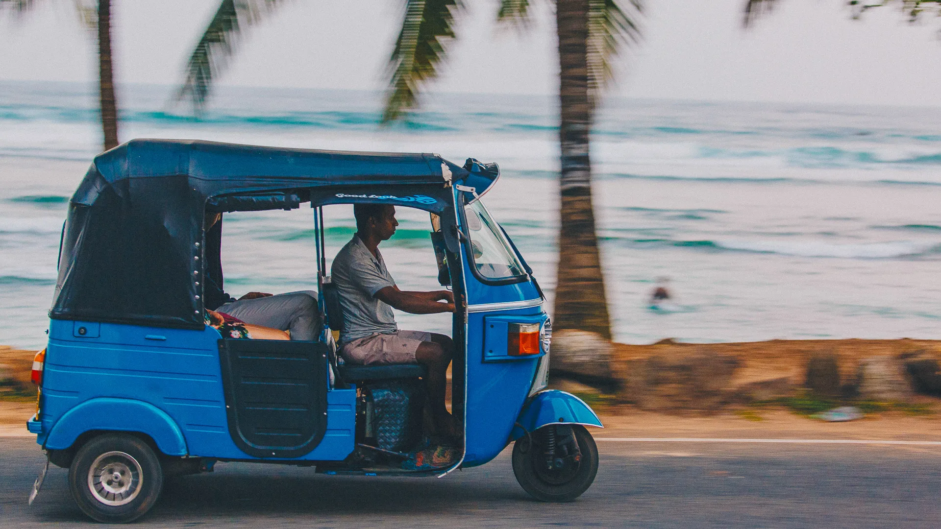Man driving a blue tuk tuk along the coast road in Sri Lanka with ocean waves in the background
