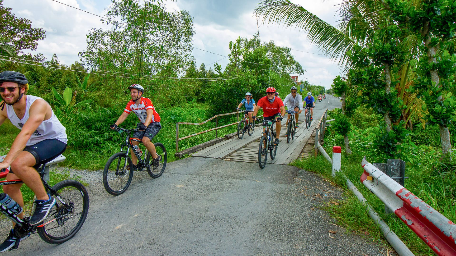 Cycling group crossing narrow rural bridge into lush palm-lined countryside in tropical Vietnam