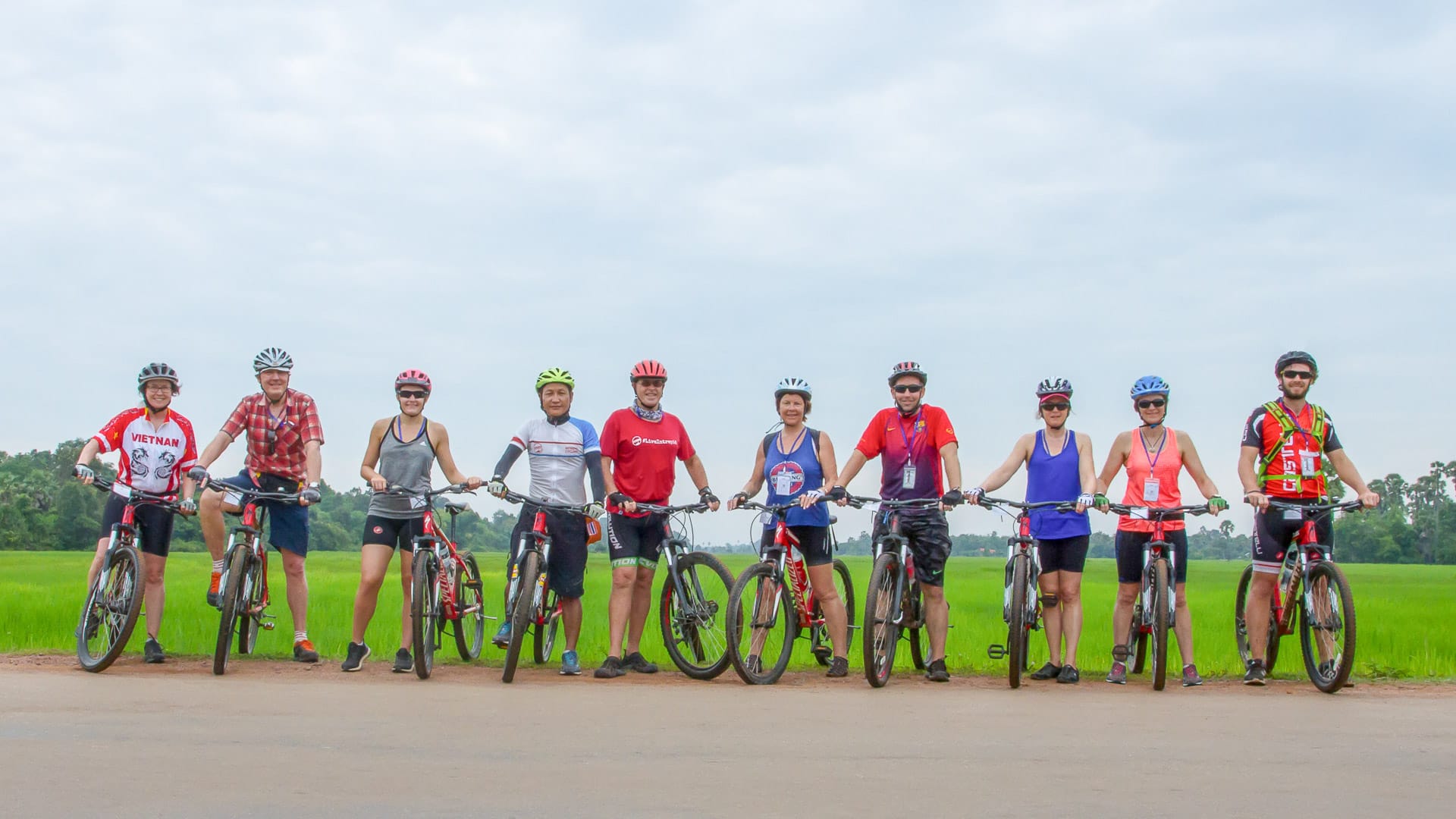 Cycling group lined up on rural road with bright jerseys and bikes beside lush rice fields