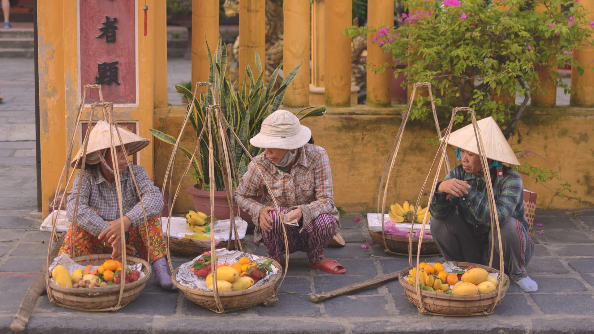 Three women in conical hats selling fruit in Hoi An, Vietnam with baskets full of mangoes and dragon fruit