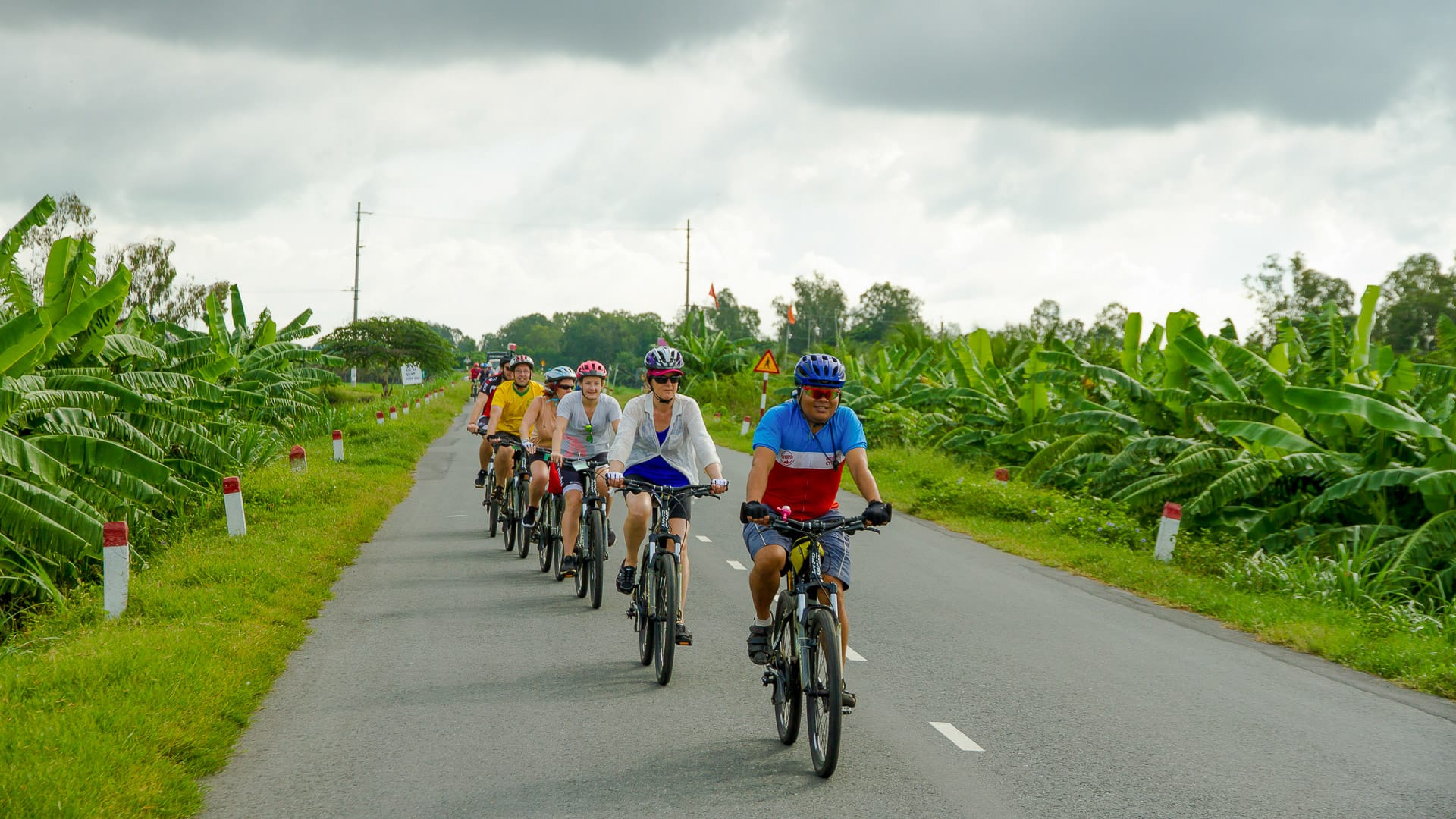 Cyclists in single file ride past vibrant banana fields under an overcast sky in rural Vietnam