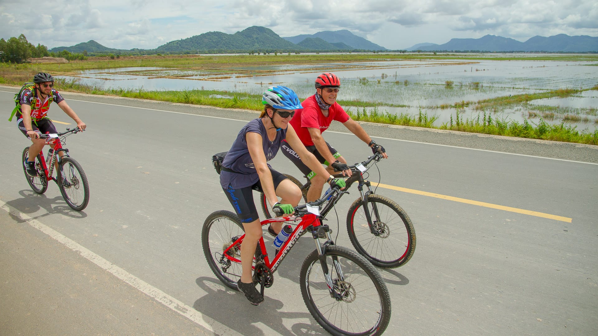 Cyclists riding beside flooded rice paddies with distant green hills in rural Vietnam