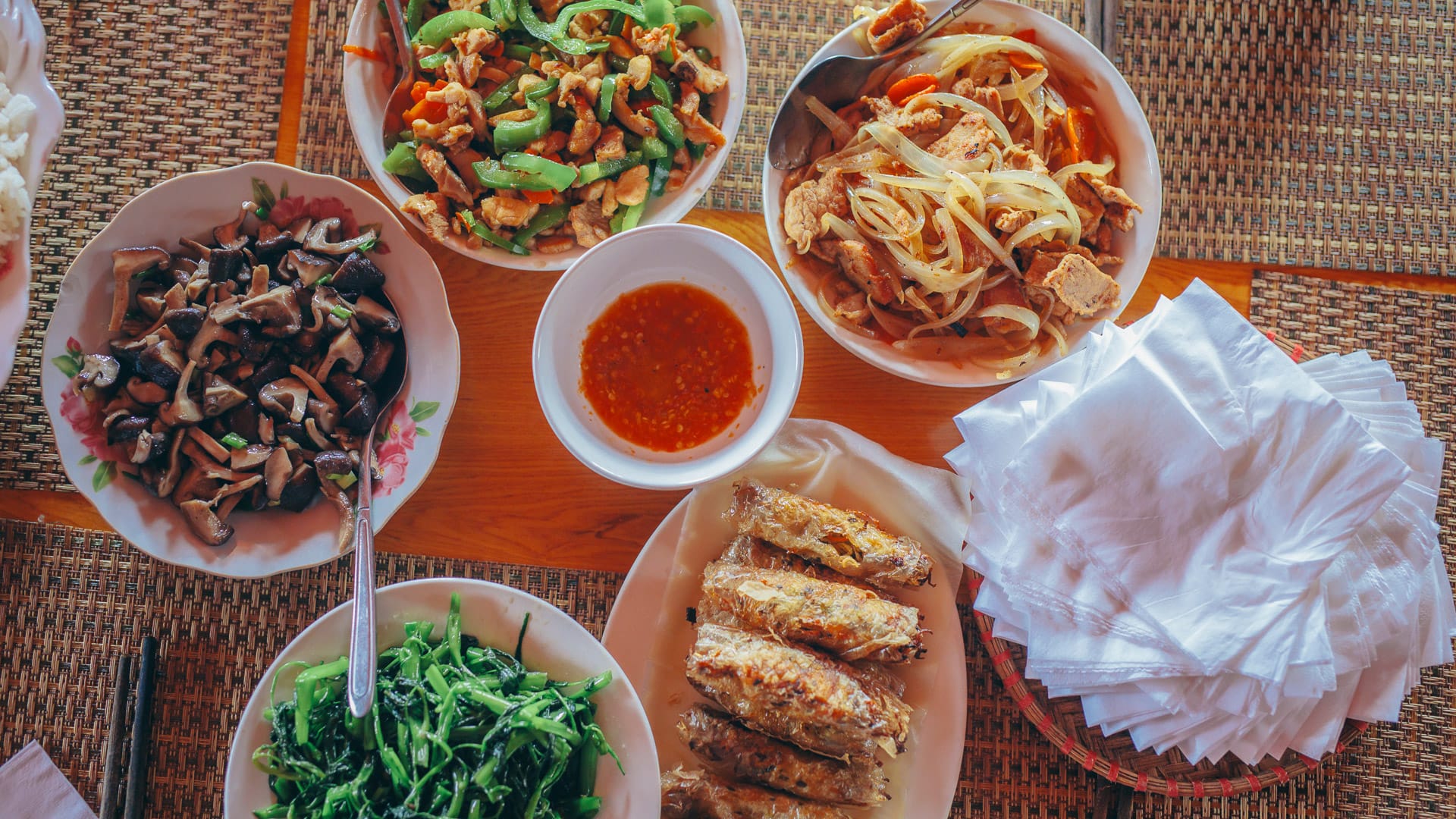 Vietnamese dishes with spring rolls, stir-fried greens, noodles and dipping sauce on woven mat table setting