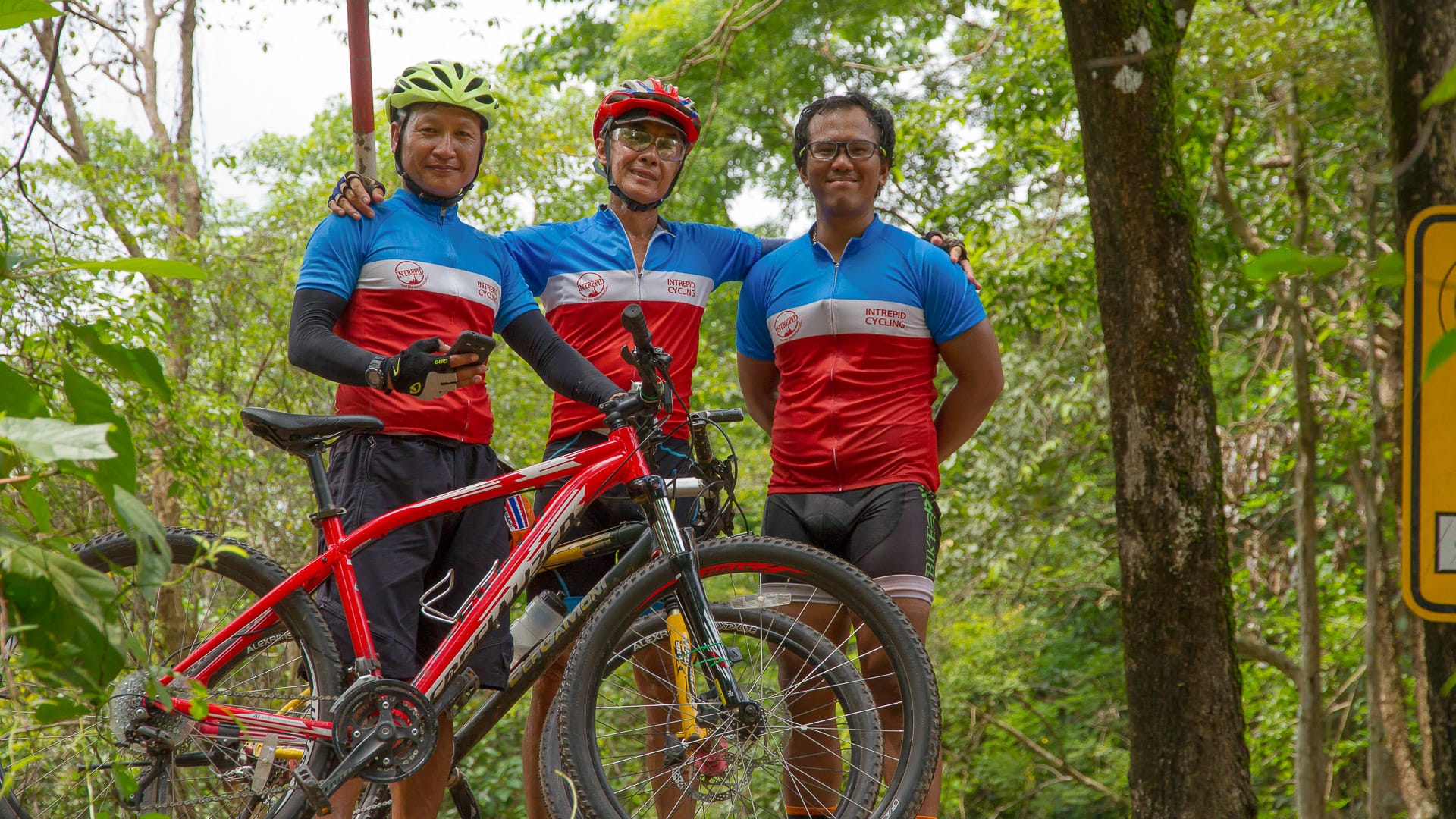 Three cyclists in matching jerseys pose with a red mountain bike in the forest