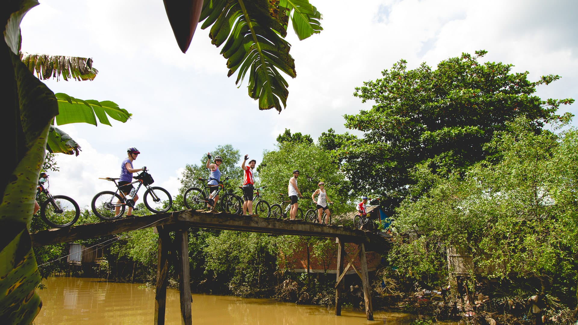 Cyclists crossing narrow wooden bridge over river in Vietnam surrounded by banana trees and lush greenery