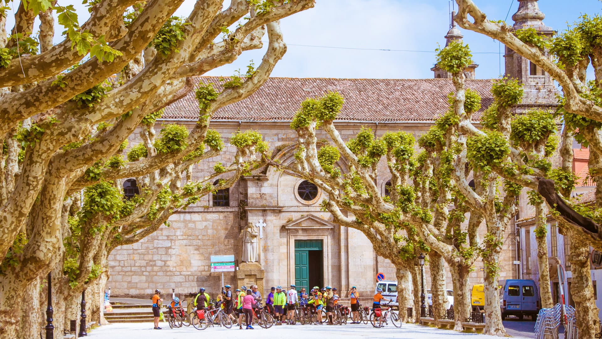 Cyclists gathered in a tree-lined plaza in front of a historic stone church with green door