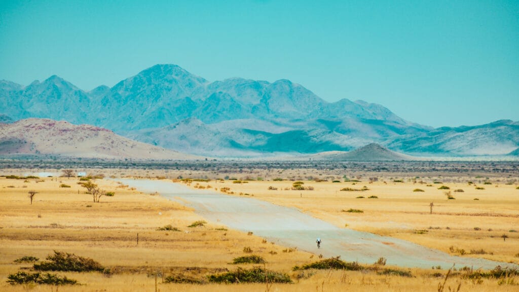 Cyclist riding along a remote dirt road through golden grasslands with hazy blue mountains rising in the distance
