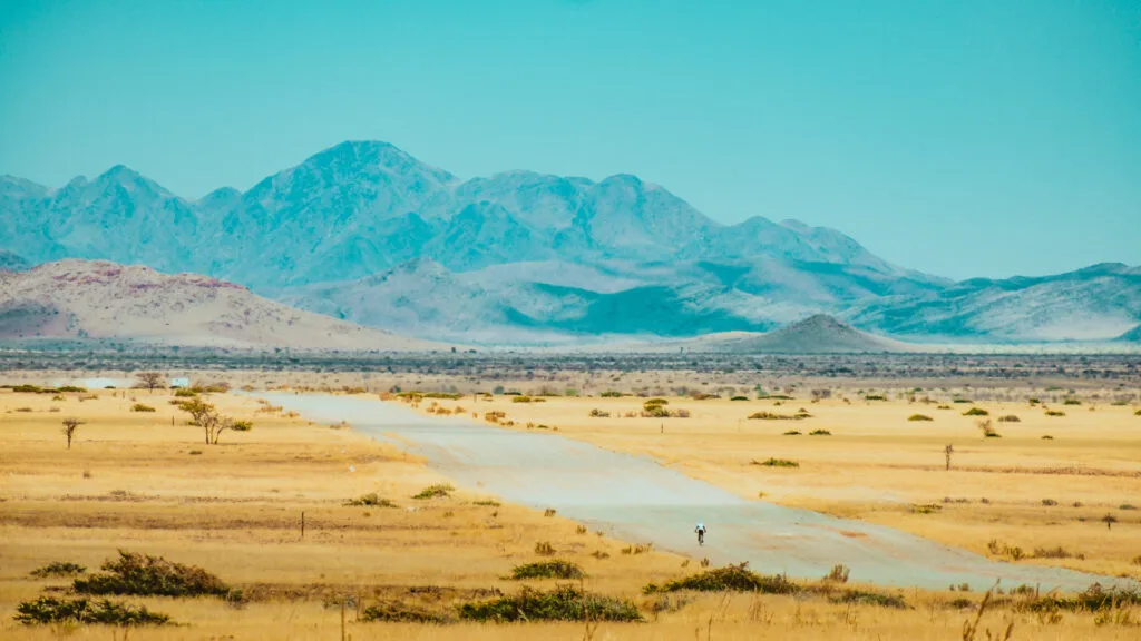 Cyclist riding along a remote dirt road through golden grasslands with hazy blue mountains rising in the distance