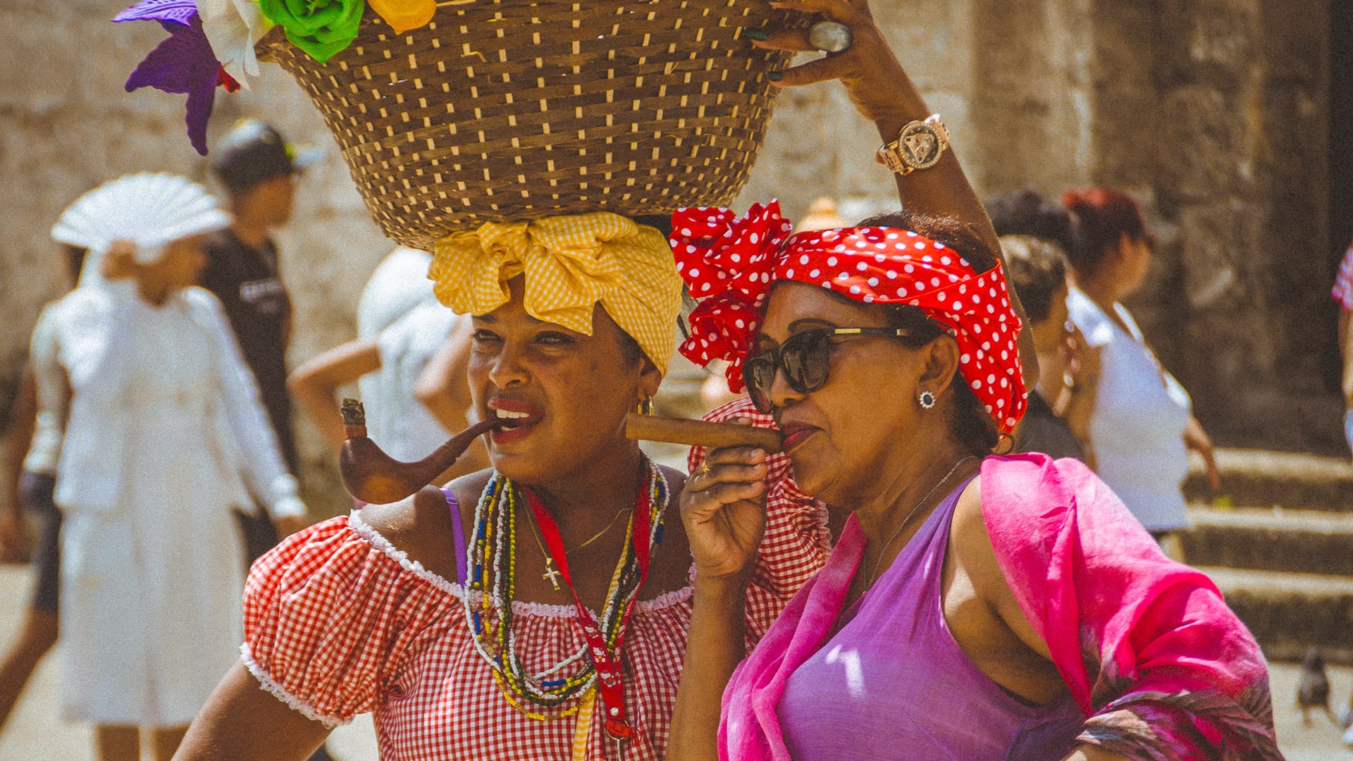 Two Cuban women in colourful traditional dress posing with cigars and a fruit basket
