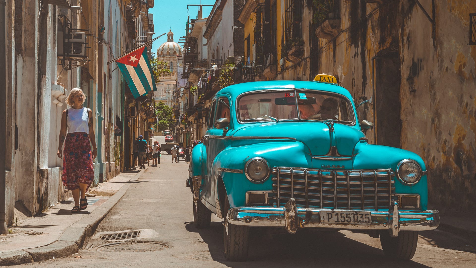 Bright blue classic taxi driving through narrow Havana street with Cuban flag flying