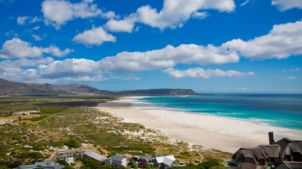 Aerial view of Noordhoek Beach in Cape Town, South Africa, with turquoise water, white sand, and mountain backdrop