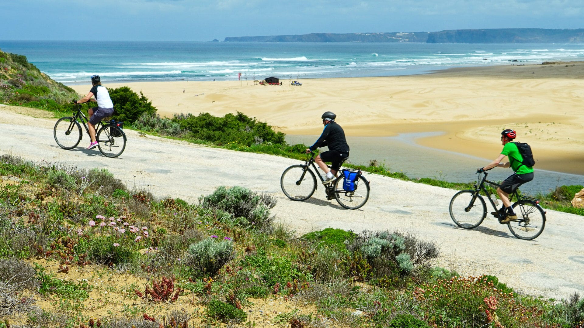Three cyclists riding along a coastal trail above a sandy beach and ocean in Portugal