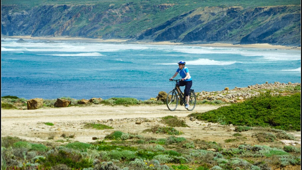Cyclist riding along a rugged coastal trail with waves crashing below and cliffs in the background