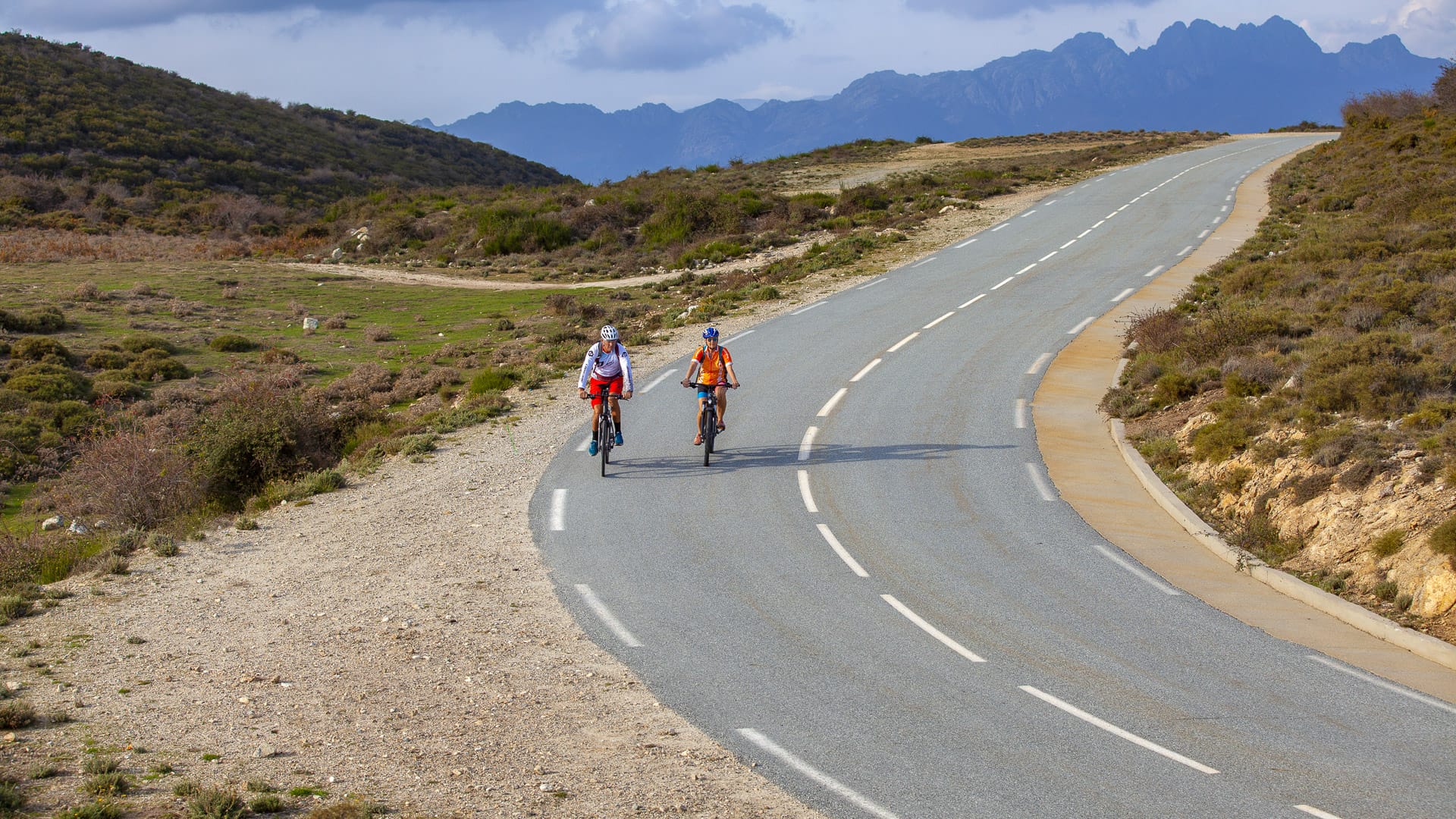 Two cyclists riding on a winding mountain road with views of Corsica’s rugged peaks