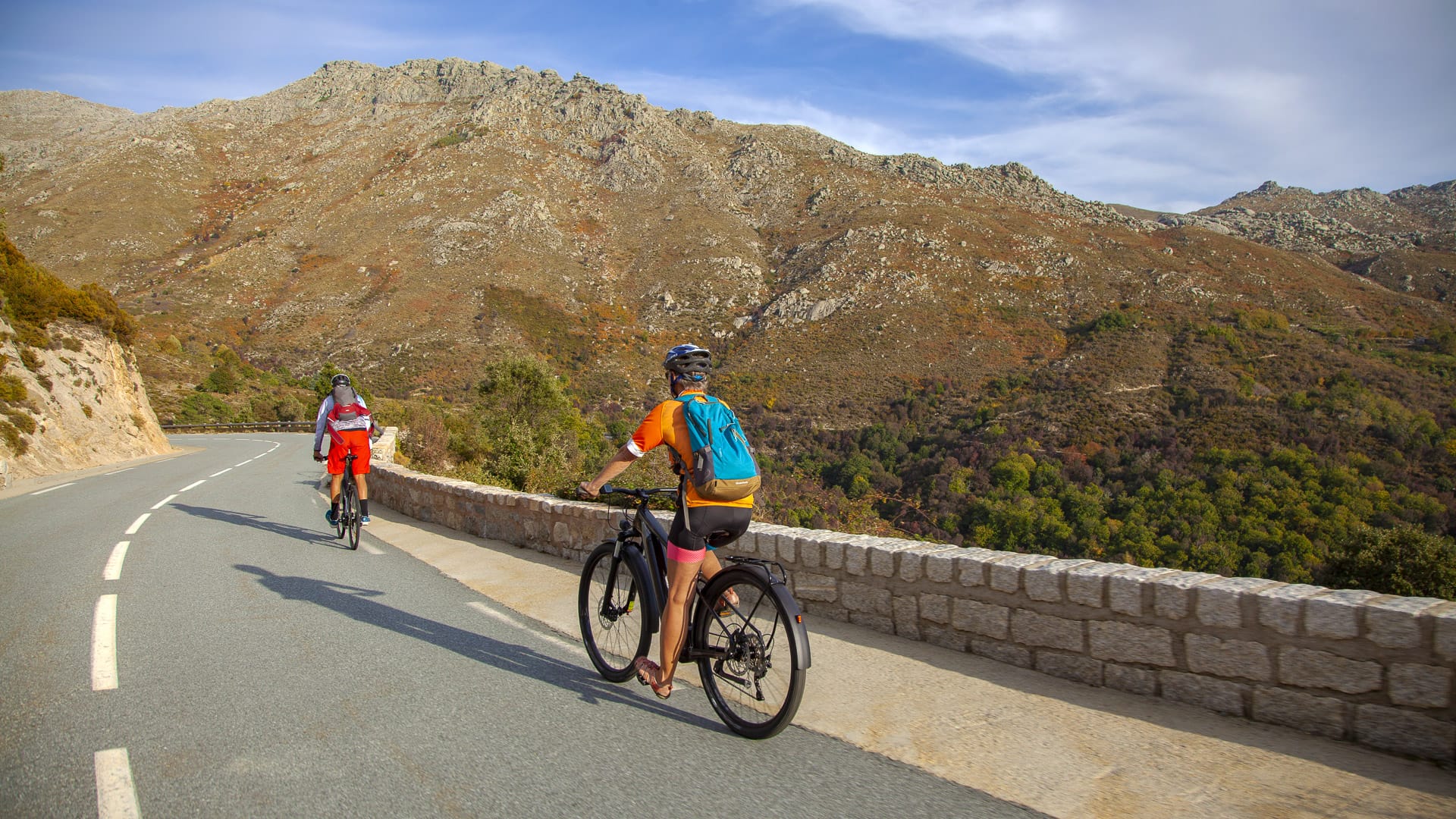 Two cyclists on a winding road with rocky slopes and forested hills in Corsica
