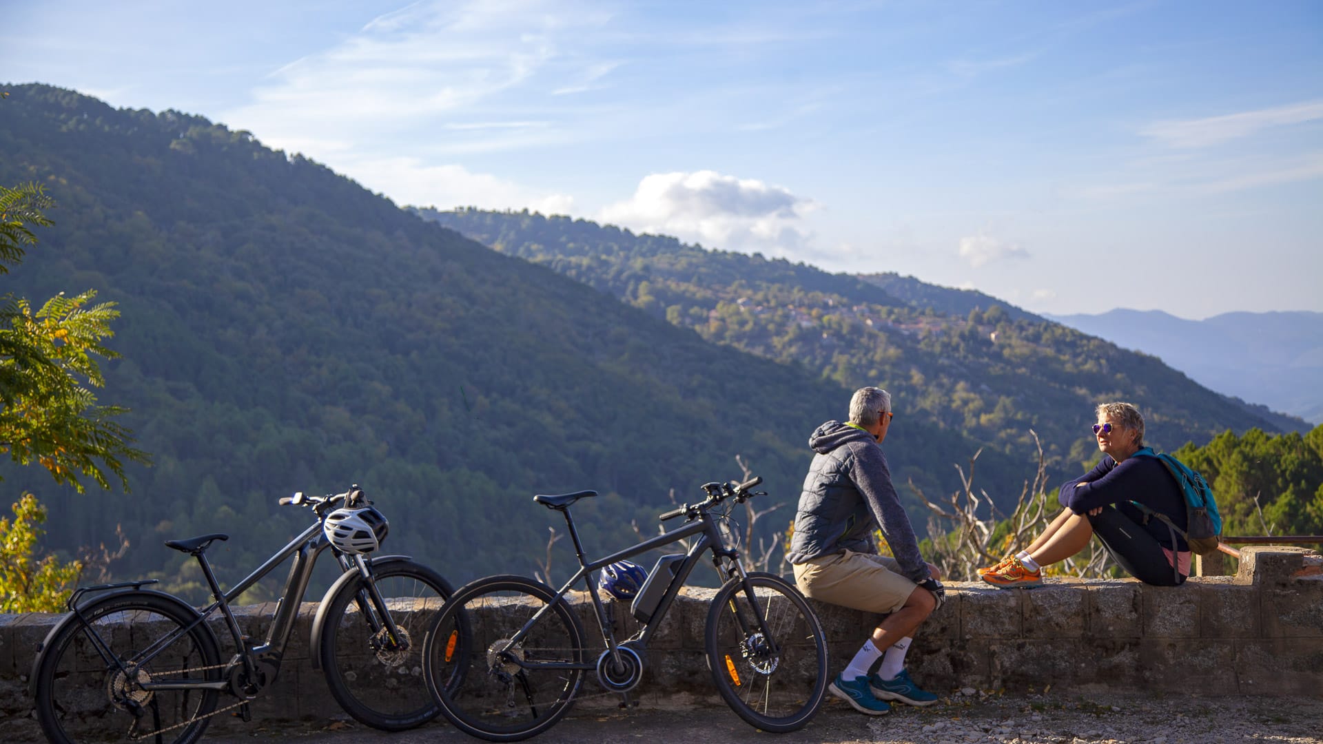 Two cyclists resting on a stone wall with a panoramic view over Corsican forest hills