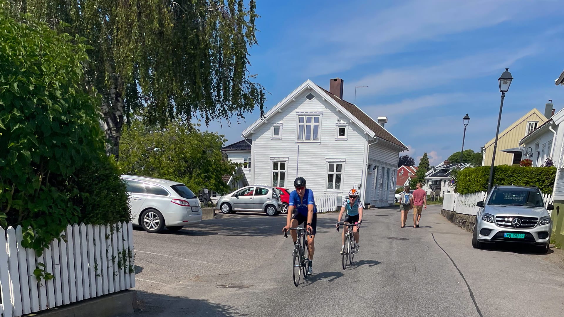 Two cyclists ride past white wooden houses in Lillesand, Norway, on a sunny summer day