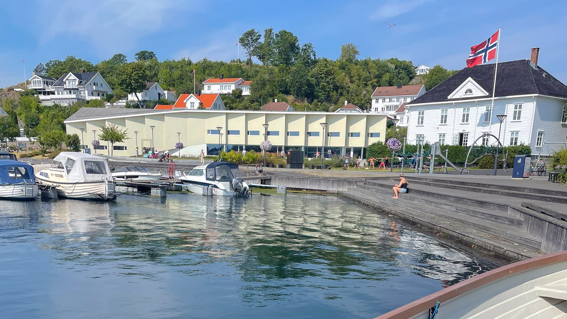 Boats moored by steps and modern library hall at Grimstad harbour with Norwegian flag flying
