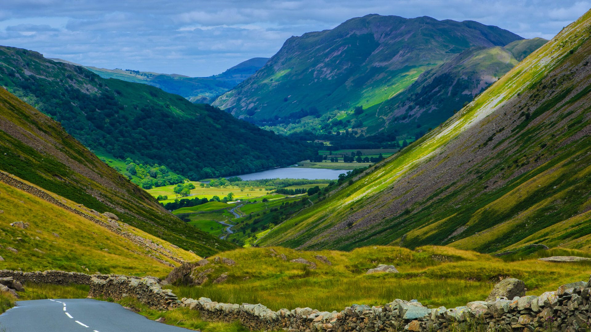 Winding road through lush green valleys in the Lake District, UK, with a lake and mountains in the distance