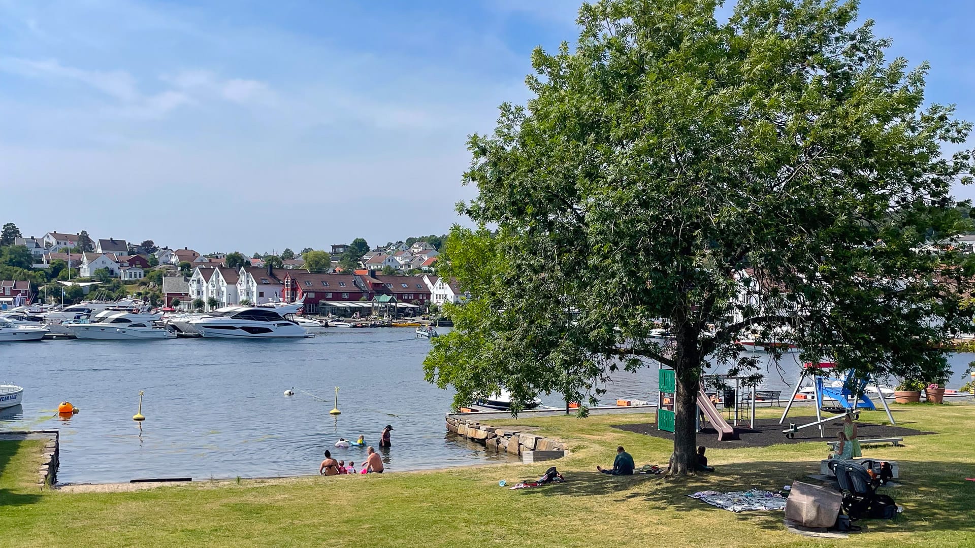 Families relax and swim by the grassy park and harbourfront in the coastal town of Lillesand, Norway