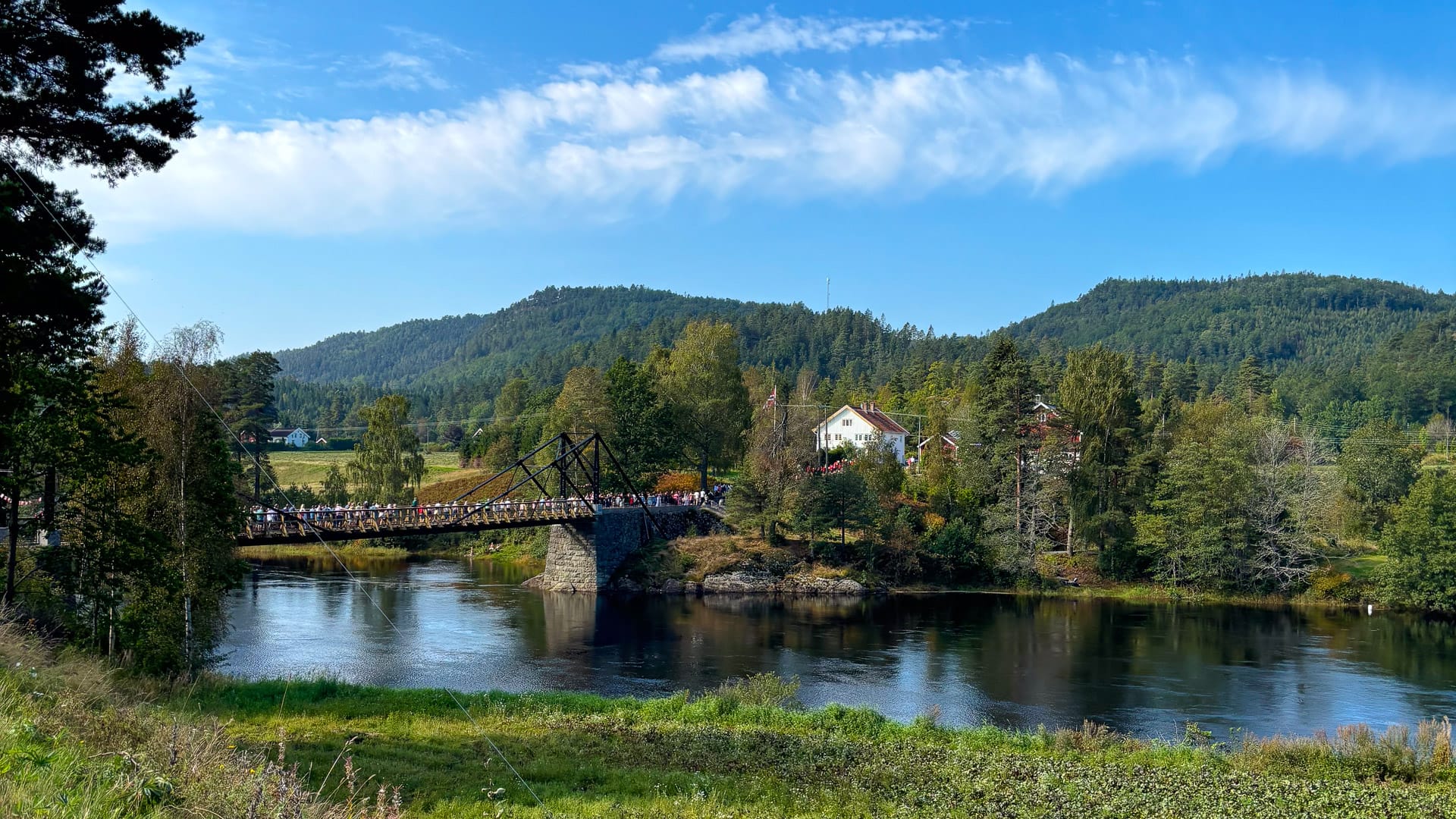 Scenic view of Rugsland bridge crossing a calm river near Birkeland, surrounded by forested hills and traditional houses