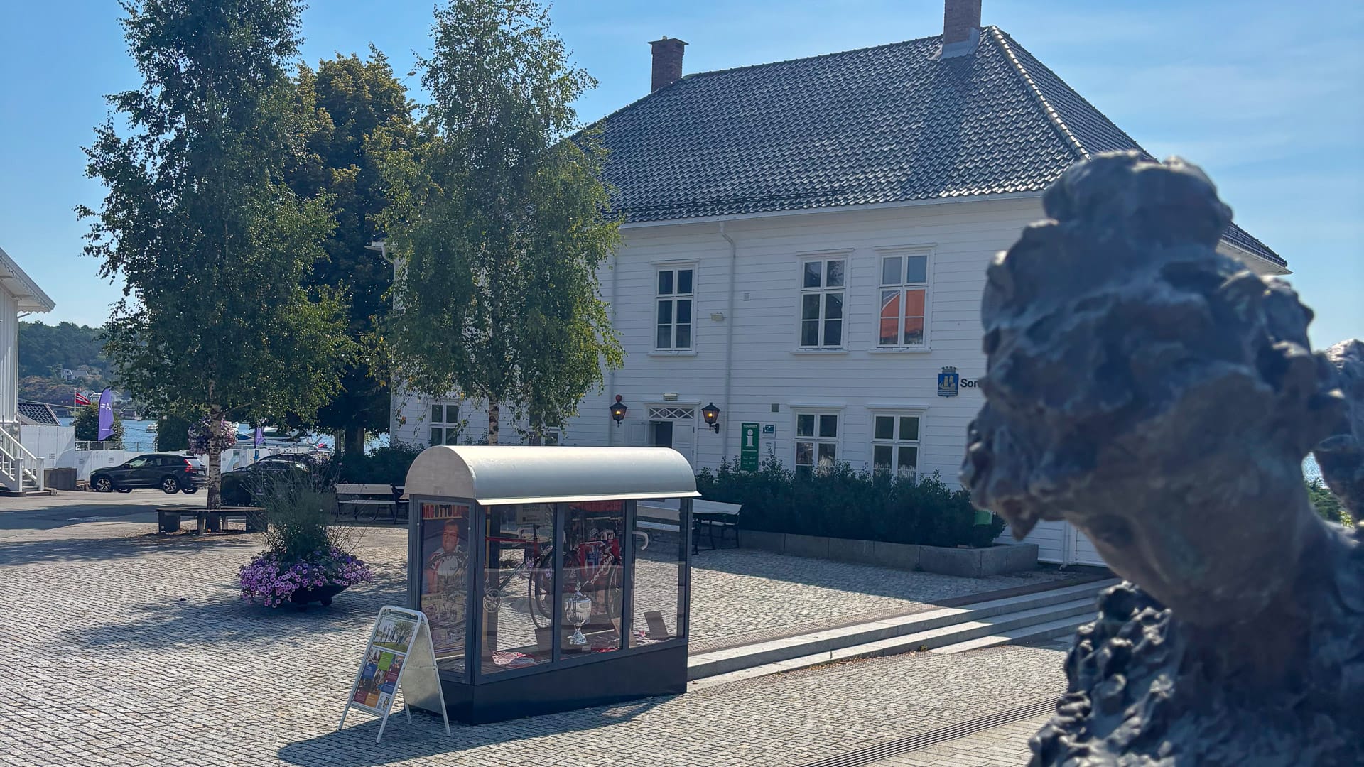 White building with trees and statue at Thor Hushovd Square in Grimstad town centre
