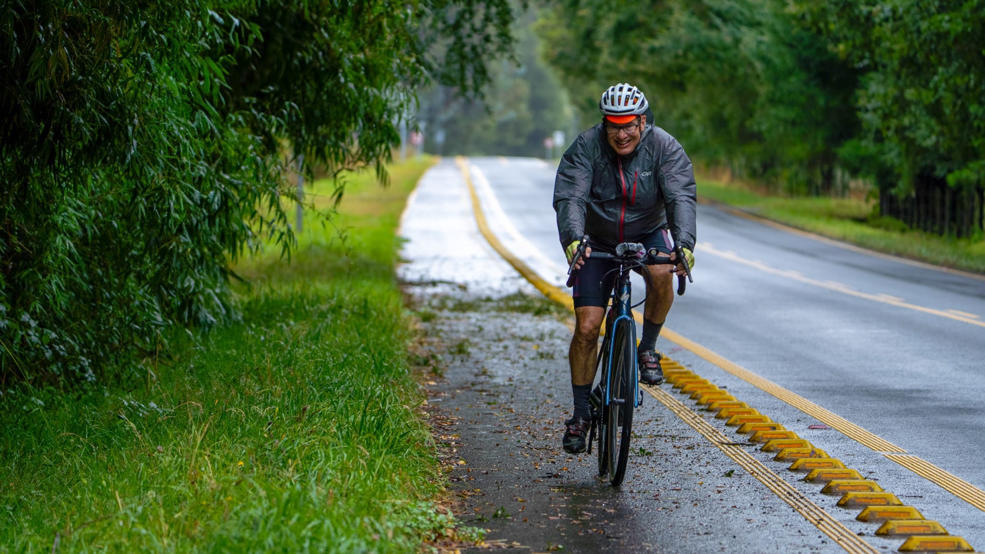 Cyclist rides wet roadside bike lane surrounded by lush greenery after rainfall