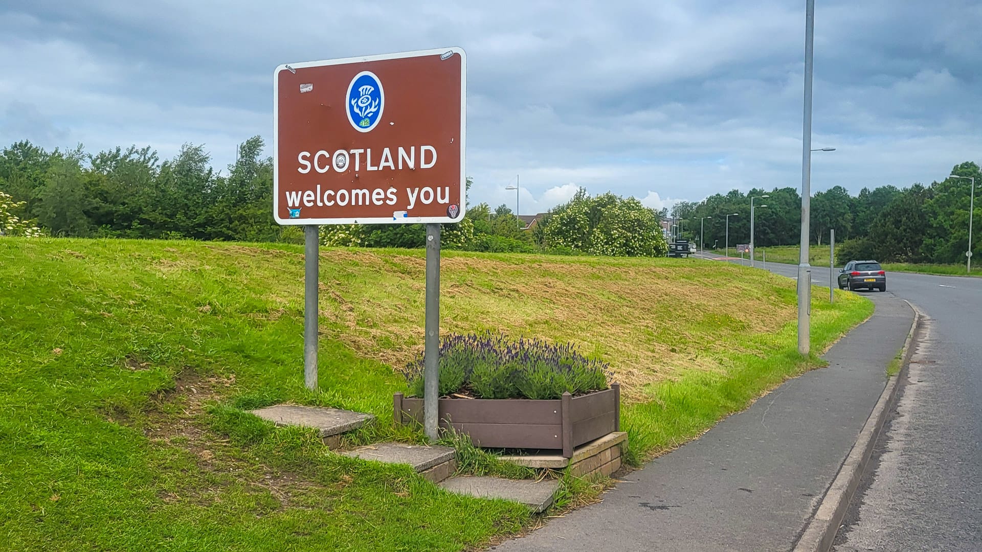 Scotland border sign reading “Scotland welcomes you” beside a road under a cloudy sky