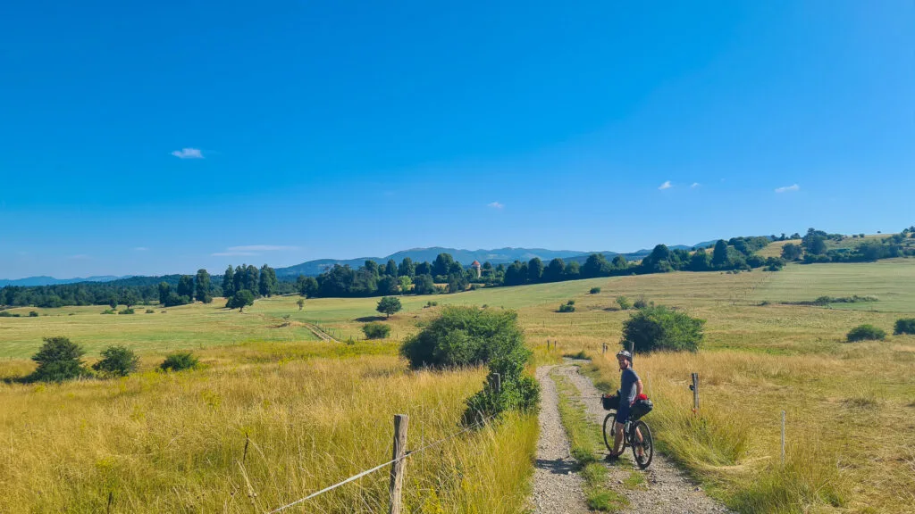 Bikepacker rides gravel trail through golden summer fields with mountain views in Slovenia