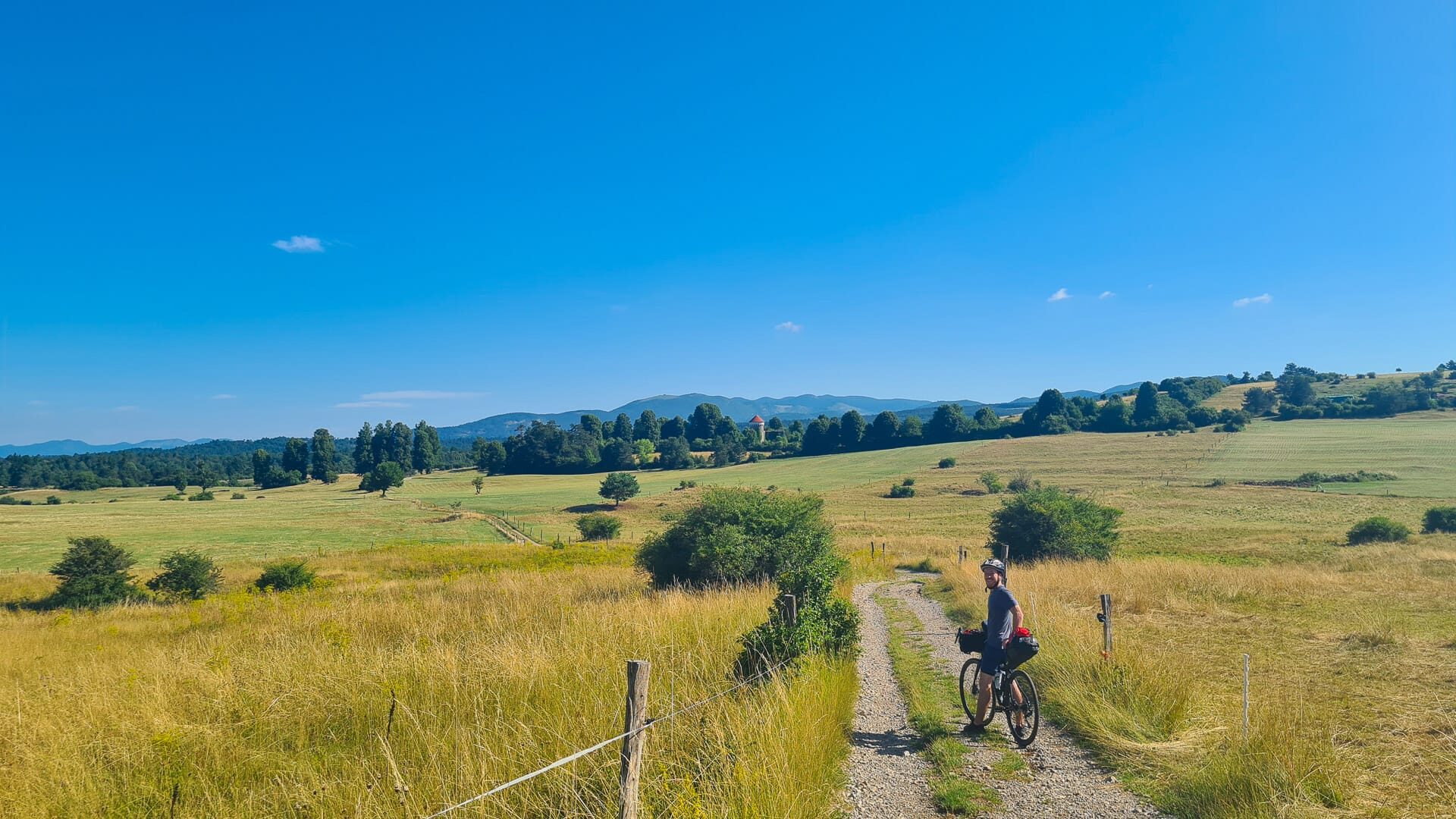 Bikepacker rides gravel trail through golden summer fields with mountain views in Slovenia