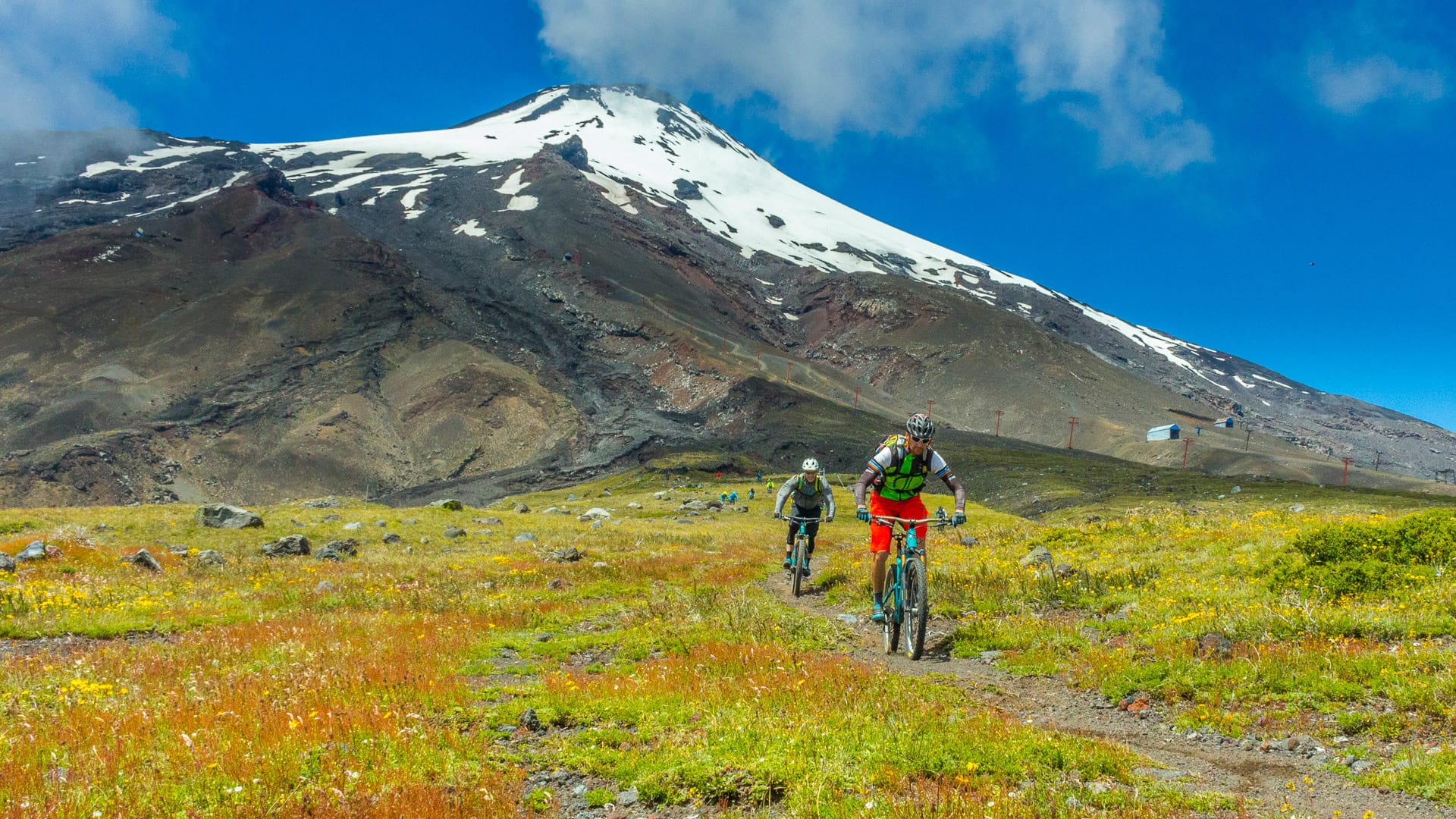 Mountain bikers ride across vibrant spring meadow towards snow-capped volcano near Pucón, Chile