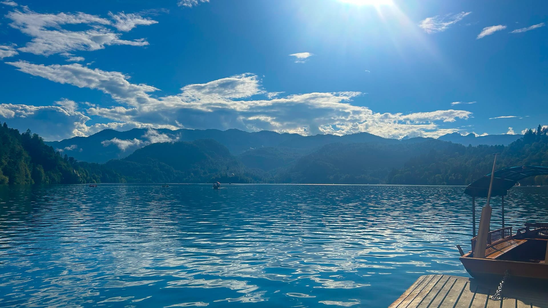 Sunlit view across Lake Bled with wooden pier and mountains in the distance