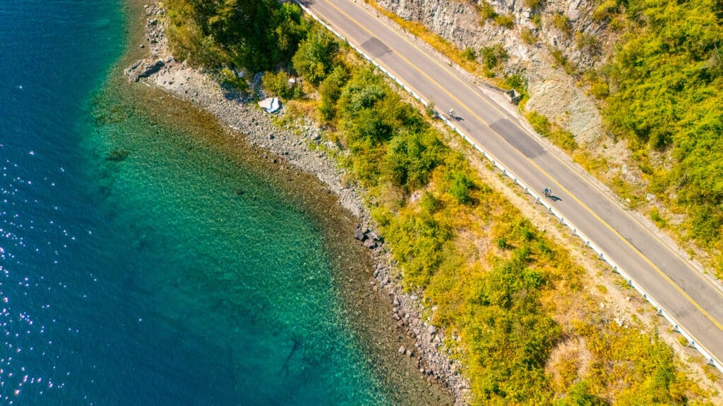Cyclists ride along coastal road beside turquoise waters and rocky shoreline