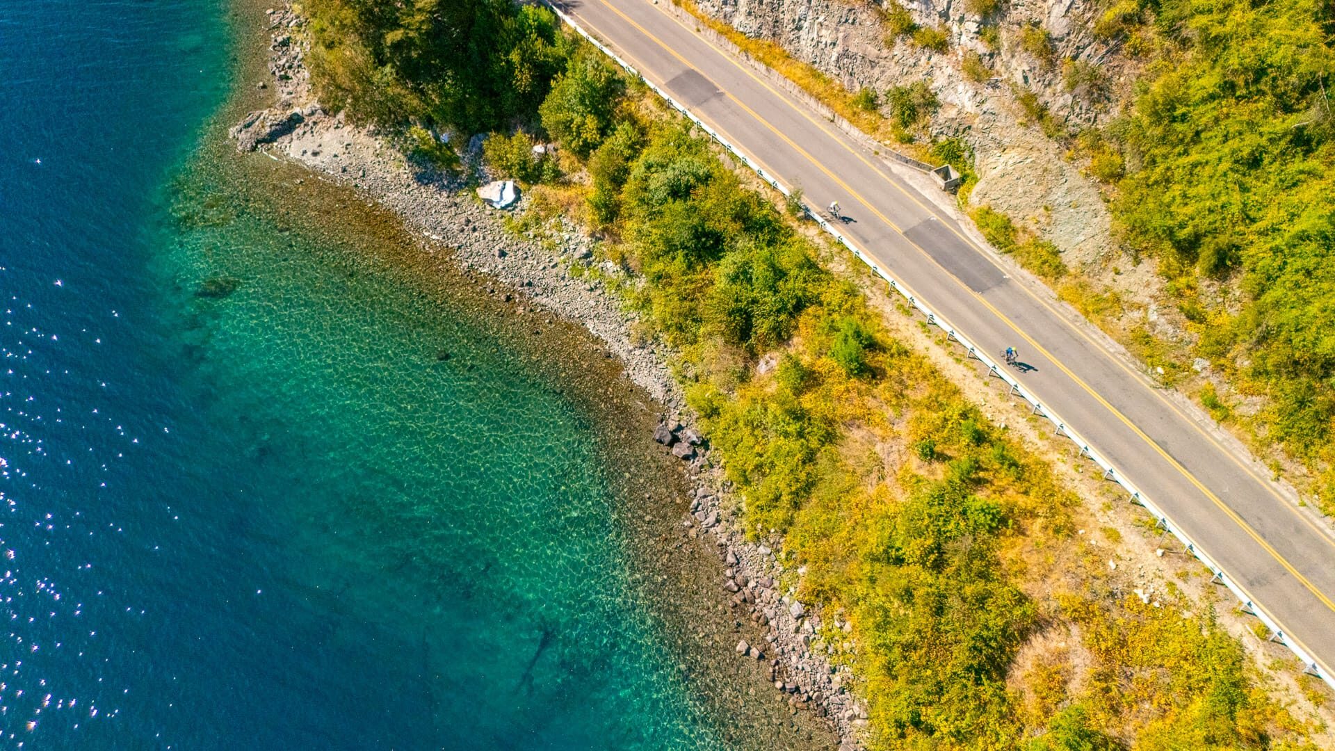 Cyclists ride along coastal road beside turquoise waters and rocky shoreline