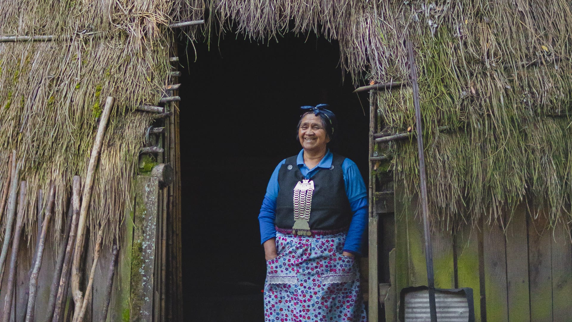 Mapuche woman smiles while welcoming visitors at the entrance of a traditional thatched ruka