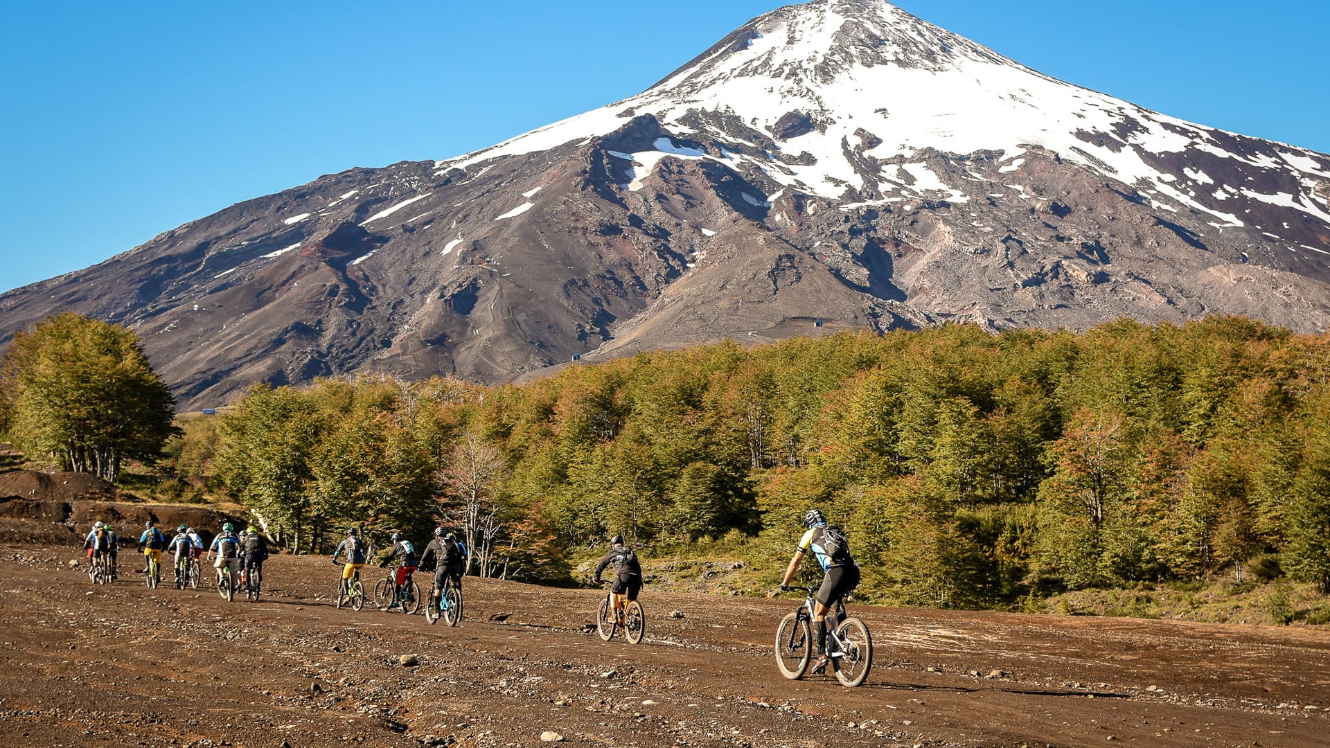 Group of cyclists ride across open terrain towards towering snow-capped volcano in Chilean landscape