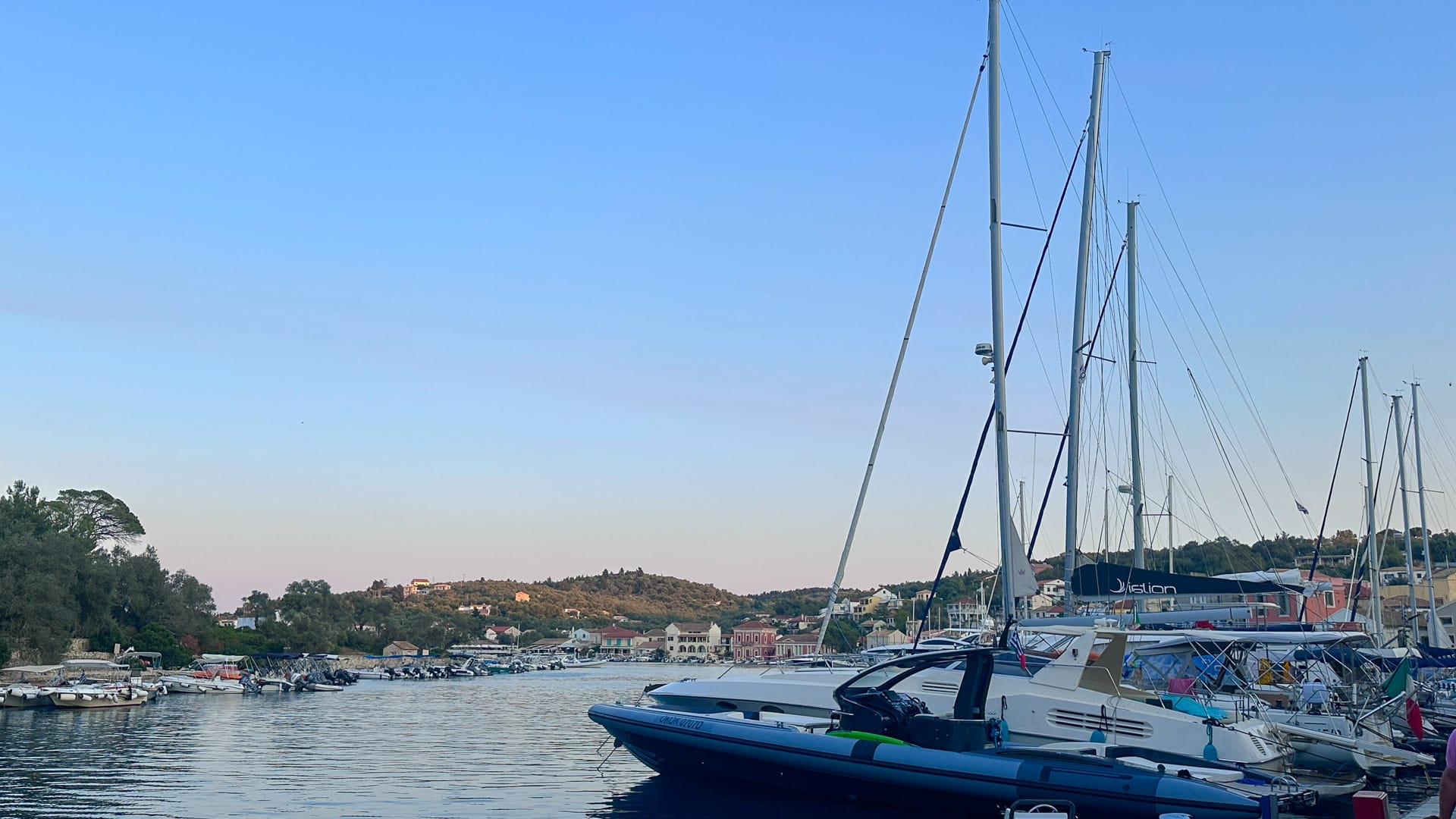 Sailboats docked in a calm marina with coastal village and hills in the background during suns