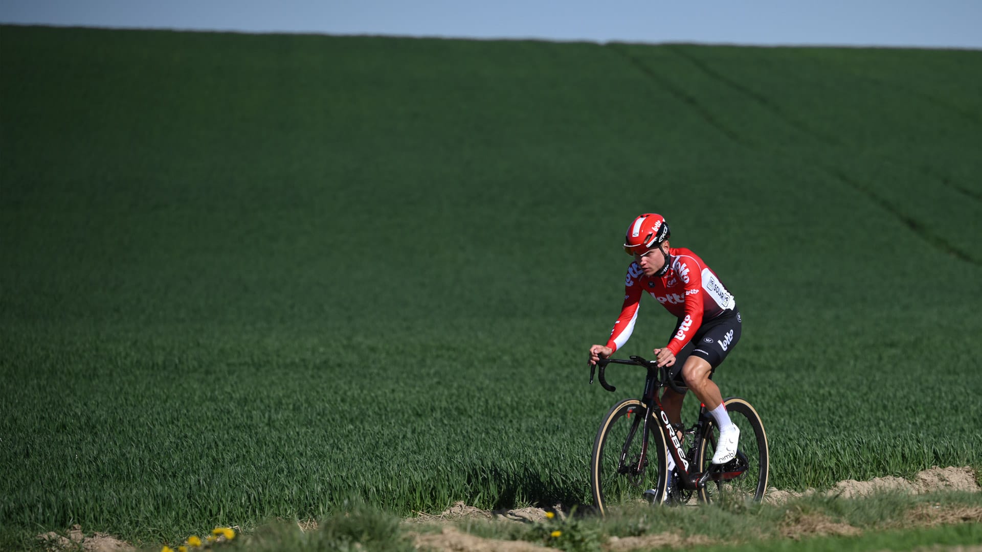 Cyclist in red Lotto Dstny kit riding beside lush green farmland near Hoegaarden, Flanders