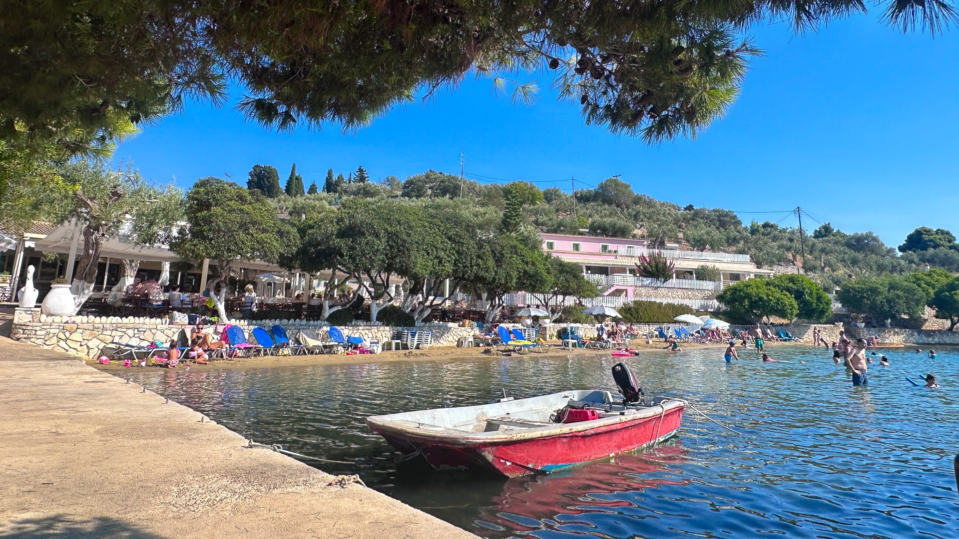 People relaxing on a beachside with sun loungers, clear shallow water, and a small red motorboat docked near the shore