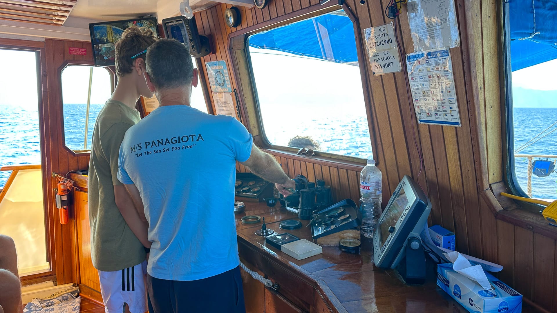 Two people at the wooden helm of a boat steering out at sea in Greece